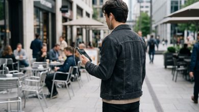 شاب يستخدم سماعات أذن بيضاء سلكية متصلة بهاتف ذكي في أحد الأماكن العصرية، في صورة تعكس عودة السماعات السلكية إلى الانتشار بين الشباب باعتبارها صيحة جديدة في عالم الموضة والتكنولوجيا.
