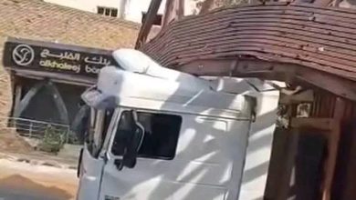 "شاحنة نقل بيضاء عالقة تحت جسر مشاة خشبي منهار جزئياً، بالقرب من لافتة بنك الخليج في منطقة حضرية."