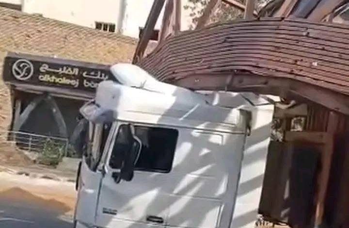"شاحنة نقل بيضاء عالقة تحت جسر مشاة خشبي منهار جزئياً، بالقرب من لافتة بنك الخليج في منطقة حضرية."