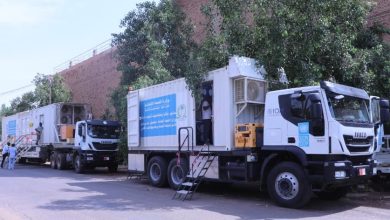 عيادة صحية متنقلة بيضاء تحمل شعار الأمم المتحدة الإنمائي (UNDP) تسير على طريق ترابي في منطقة سكنية بالخرطوم، السودان، لتقديم الخدمات الطبية.