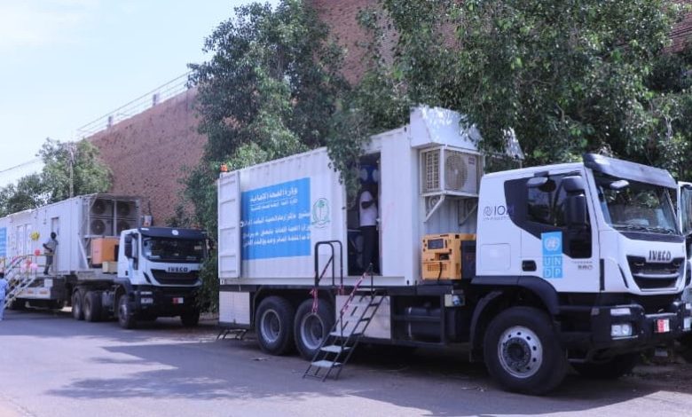 عيادة صحية متنقلة بيضاء تحمل شعار الأمم المتحدة الإنمائي (UNDP) تسير على طريق ترابي في منطقة سكنية بالخرطوم، السودان، لتقديم الخدمات الطبية.