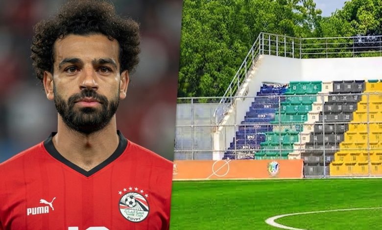 محمد صلاح بقميص منتخب مصر بجانب ملعب جوبا في جنوب السودان، مع ظهور أرضية العشب الصناعي ومدرجات الاستاد، في صورة رياضية تعبر عن غياب قائد الفراعنة عن مواجهة جنوب السودان بسبب طبيعة أرضية الملعب
